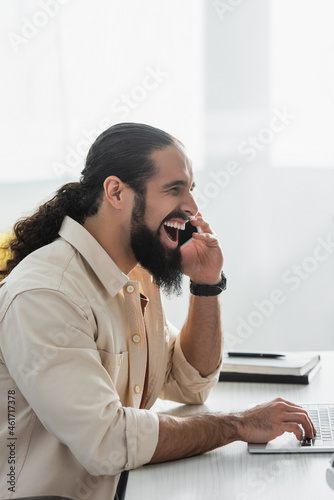 excited hispanic freelancer laughing while talking on cellphone near laptop