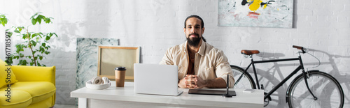 bearded latin freelancer sitting near laptop and graphic tablet in home studio, banner photo