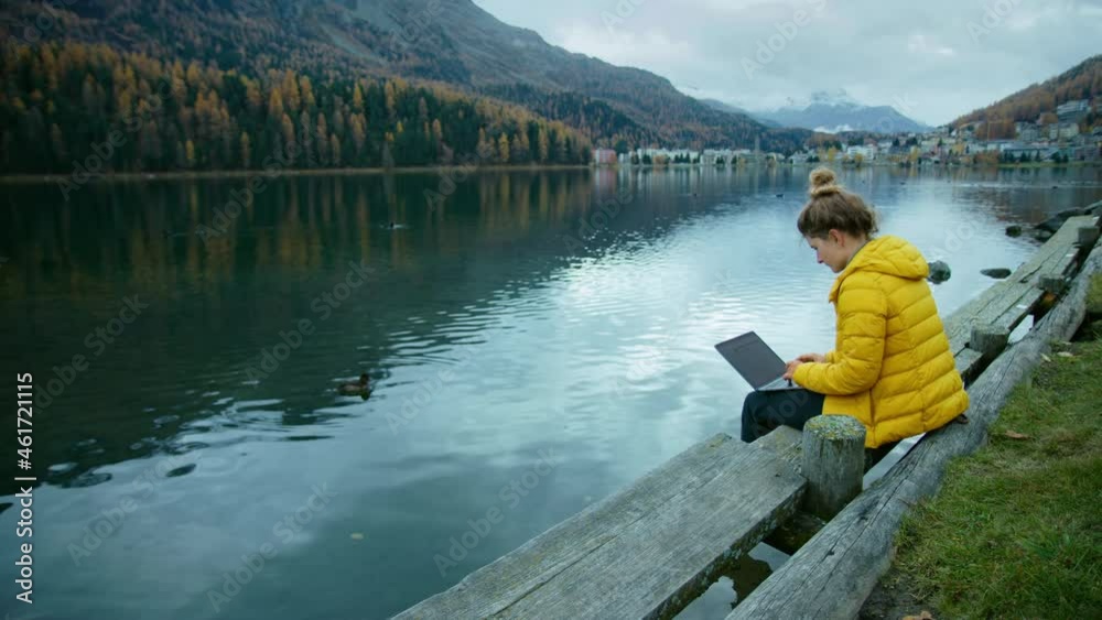 Young woman in bright yellow jacket work on side of lake on her laptop. Female freelancer work remotely during vacation. Cinematic modern millennial work style concept Young woman in bright yellow jacket work on side of lake on her laptop. Female freelancer work remotely during vacation. Cinematic modern millennial work style concept