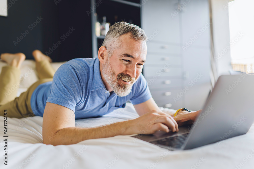 nice mature man at hotel with grey beard relaxing on bed with laptop nice mature man at hotel with grey beard relaxing on bed with laptop