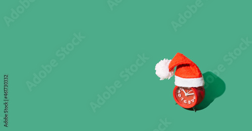 red alarm clock in red Christmas cap with white pompon on green background, koncept new year and christmas, fashionable minimalism, free space for text