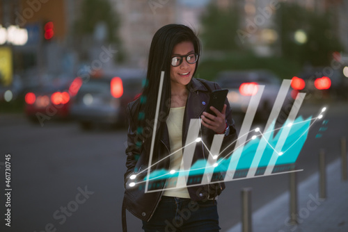 A young beautiful Asian woman is monitoring graphical and statistical data from her phone device, depicted as a holograph around her, while in the city street