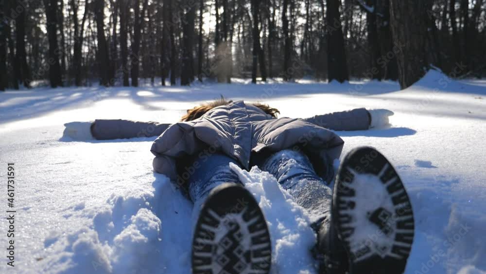 Young girl in winter clothes lying on snowy meadow and making angel. Happy woman playing in snow enjoying winter time. Cheerful lady having fun outdoor. Concept of winter holiday. Slow motion Close up