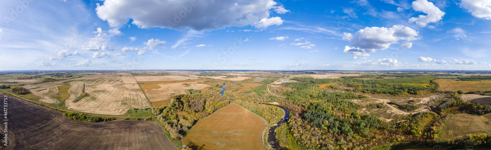 Aerial panorama of a vast prairie in autumn colors with white clouds in a blue sky. A river cuts through the scene.
