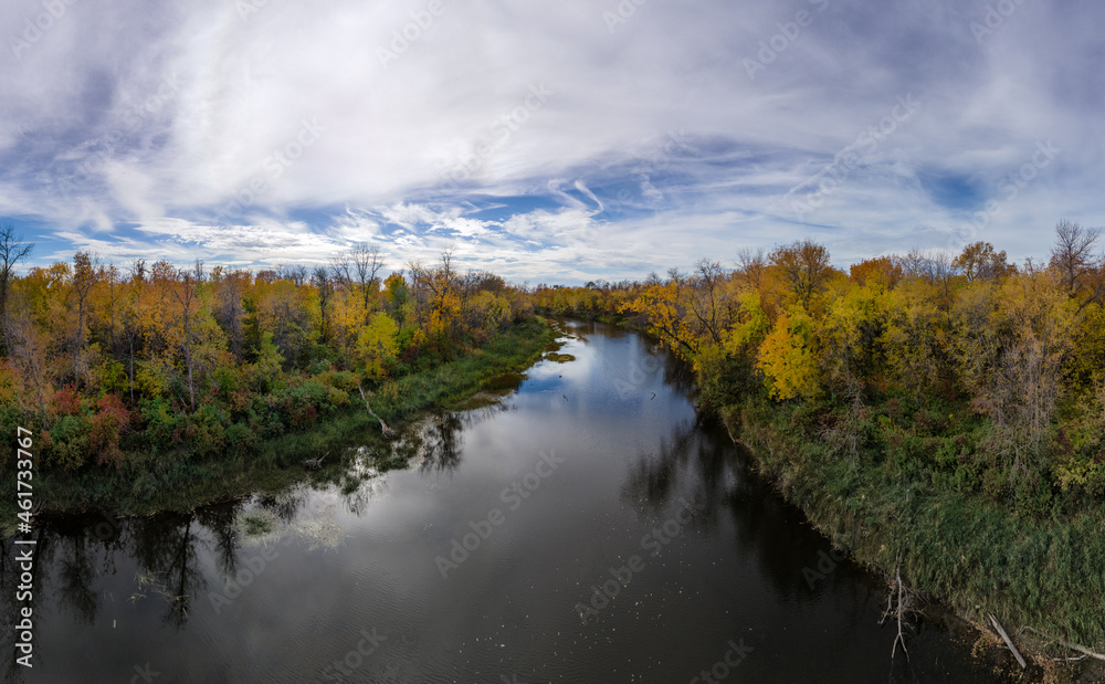 Aerial view of brightly colored autumn trees surrounding a large calm river and a sky full of white clouds with blue patches.

