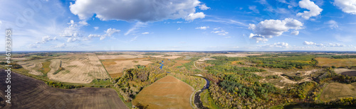 Aerial panorama of a vast prairie in autumn colors with white clouds in a blue sky. A river cuts through the scene. 