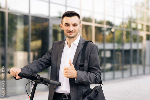 Young businessman in elegant suit standing outdoors, cheerfully smiling and showing thumb up. Handsome businessman with his electric scooter in front the business building.