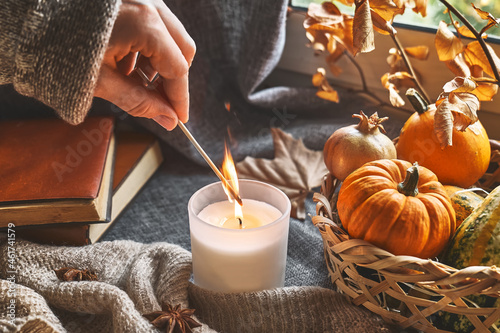 Hand with burning match lighting a candle on the windowsill with cozy autumn still life with pumpkins, knitted woolen sweater and books. Autumn home decor. Cozy fall mood. Thanksgiving. Halloween. photo