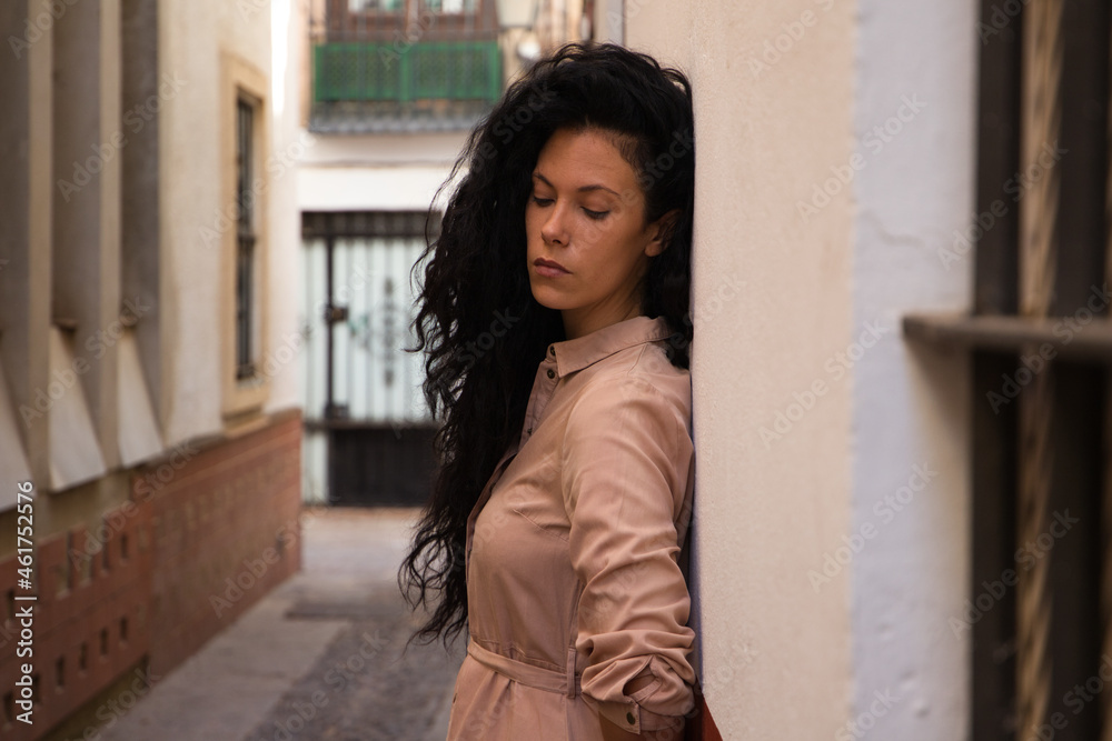 beautiful woman with dark curly hair leaning against the wall of a typical european house in a mediterranean style city. The woman is serious and sad. Concept expressions. beautiful woman with dark curly hair leaning against the wall of a typical european house in a mediterranean style city. The woman is serious and sad. Concept expressions.