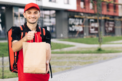 Happy young courier is proud of his job smiling standing in the street. Home delivery. Outdoor portrait of delivery man with red uniform holding food bags waiting for customer