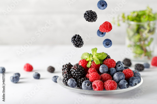 Assorted different berries with mint leaves on a glass plate on marble table. Berries falling flying