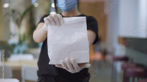 Close-up paper bag with packed lunch in female Caucasian hands with blurred young woman in coronavirus face mask at background. Unrecognizable waitress posing in restaurant with take-away order