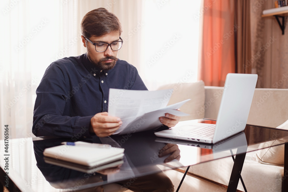 Entrepreneur in eyeglasses works with a laptop and keeps a document in a home office. Man holding paper documents, chatting online with clients on laptop at workplace. Entrepreneur in eyeglasses works with a laptop and keeps a document in a home office. Man holding paper documents, chatting online with clients on laptop at workplace.