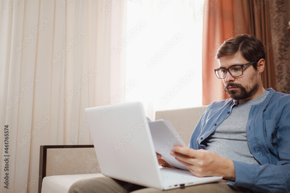 Entrepreneur in eyeglasses works with a laptop and keeps a document in a home office. Man holding paper documents, chatting online with clients on laptop at workplace. Entrepreneur in eyeglasses works with a laptop and keeps a document in a home office. Man holding paper documents, chatting online with clients on laptop at workplace.