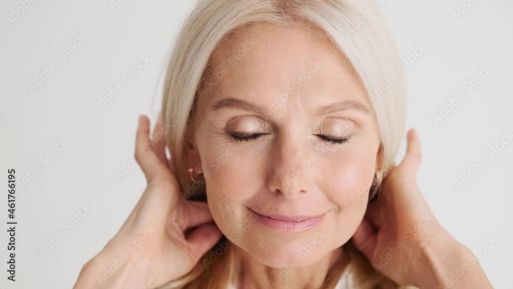 Close-up view of the smiling mature woman correcting her hairstyle at the camera as a mirror in the white studio Close-up view of the smiling mature woman correcting her hairstyle at the camera as a mirror in the white studio