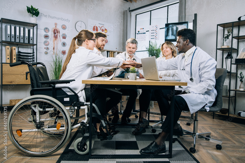Five competent multiracial doctors with one woman using wheelchair stacking their hands together during successful meeting at office. Concept of medicine, science and cooperation.