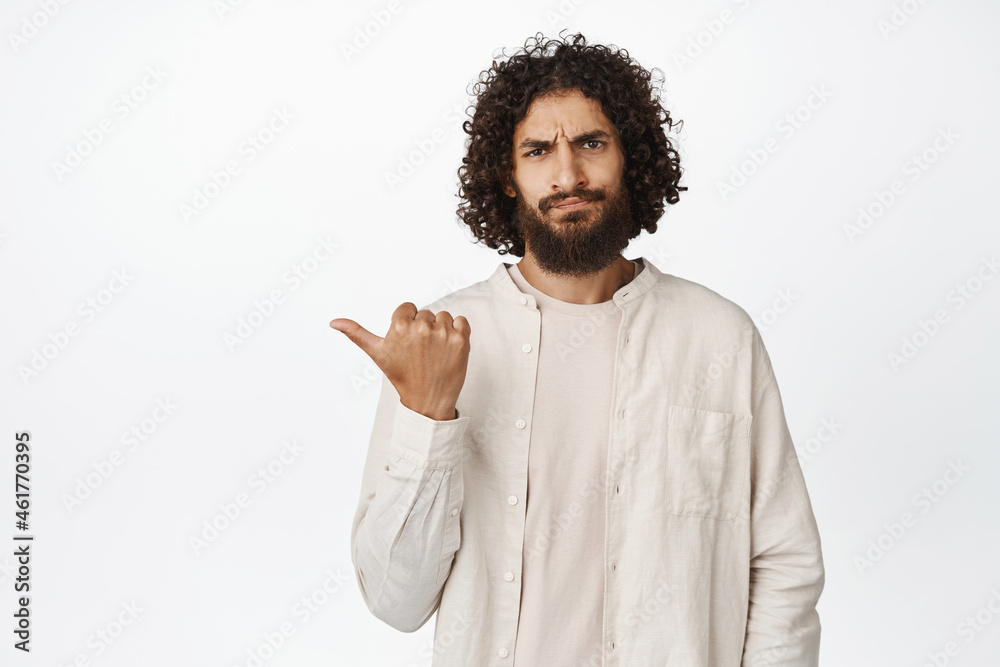 Portrait of arab guy pointing left, sulking and frowning, disapprove smth bad, dislike promo, standing over white background Portrait of arab guy pointing left, sulking and frowning, disapprove smth bad, dislike promo, standing over white background