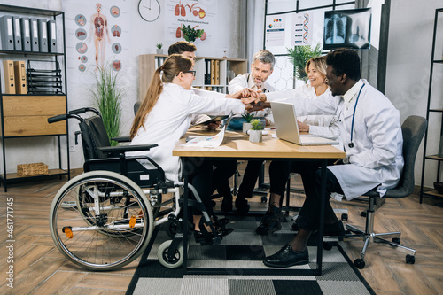 Multiracial doctors with one woman using wheelchair holding their hands together in circle during successful meeting at office. Concept of medicine, science and cooperation.