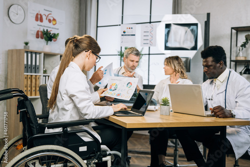 Female doctor in wheelchair working on wireless laptop during conference meeting with multiracial colleagues. Group of therapists cooperating for progress in common medical research.