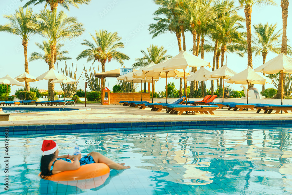 Young brunette woman in santa claus hat in a swimming circle in the pool in the Christmas holidays on the background of palm trees.