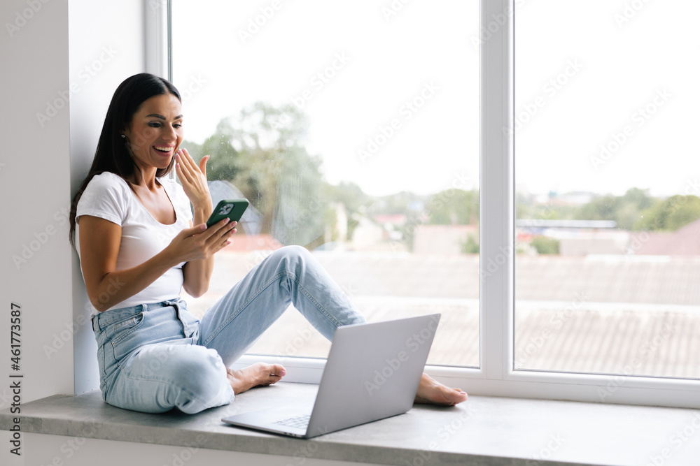 Beautiful young woman is using a mobile phone and laptop surprised on windowsill Beautiful young woman is using a mobile phone and laptop surprised on windowsill