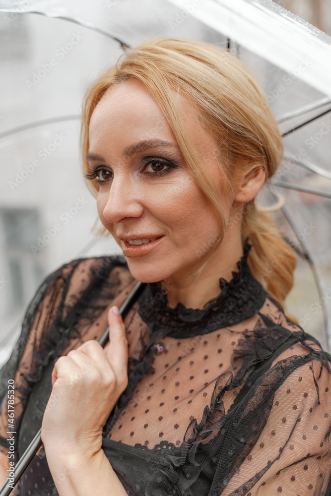 The blonde stands under a transparent umbrella during the rain. The fall season. Rear view. The woman is dressed in a black lace dress, her hair pulled back in a ponytail. The blonde stands under a transparent umbrella during the rain. The fall season. Rear view. The woman is dressed in a black lace dress, her hair pulled back in a ponytail.