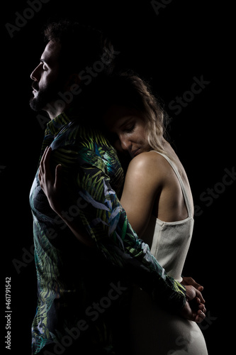 Couple posing on black background. Girl hugging boy in shadow from behind.