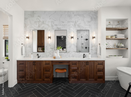 Gorgeous ensuite bathroom in luxury home. Features black herringbone tile floor  marble tile backsplash and wall  double vanity and sconce lights.