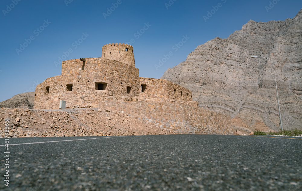 Stone walls of small medieval Arabian fort under tall mountain cliffs. Fortress in Bukha, Musandam peninsula, Oman. Hot, hazy day in Arabian desert.