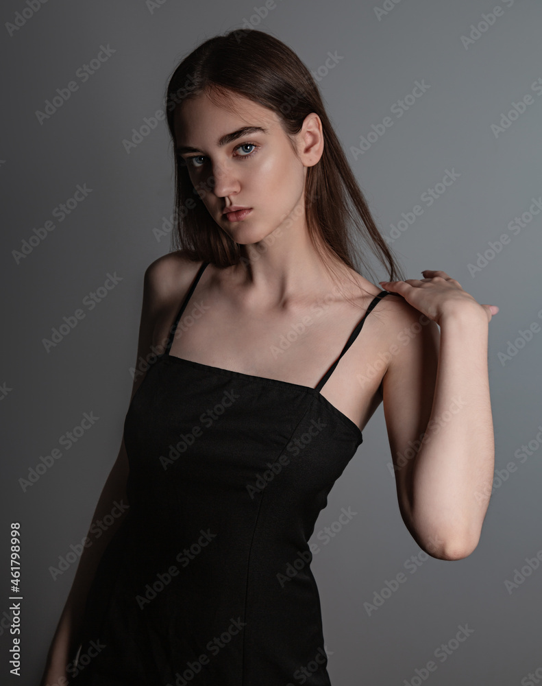 Naklejka premium Beautiful woman with nude makeup and volume brown long hair looking mystery in black dress on grey background. Closeup. Art