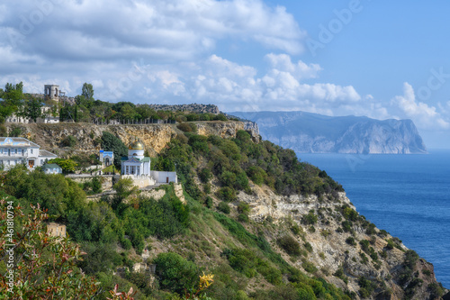View of the ancient monastery on a picturesque rock covered with green plants and the Black Sea. Mountains in the distance in the haze. Summer sunny day. Golden domes glisten in the sun. Crimea