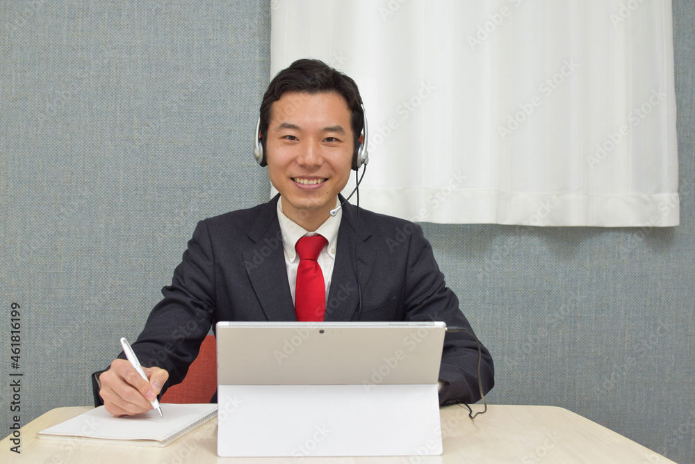 A young Asian man with a headset wearing business suit talks on video meeting app and writes a memo on a notebook with smile A young Asian man with a headset wearing business suit talks on video meeting app and writes a memo on a notebook with smile