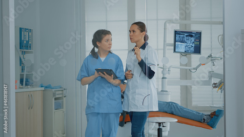 Professional dentist talking to nurse and looking at digital tablet for teeth diagnosis after examination in dental cabinet. Orthodontical team using device tool for dentistry healthcare