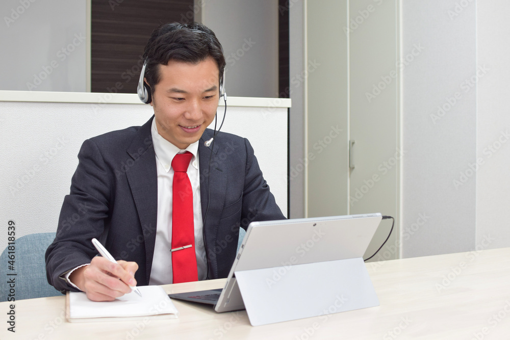 A young Asian man wearing a headset takes a memo on his notebook while talking with his customer during telework at home A young Asian man wearing a headset takes a memo on his notebook while talking with his customer during telework at home