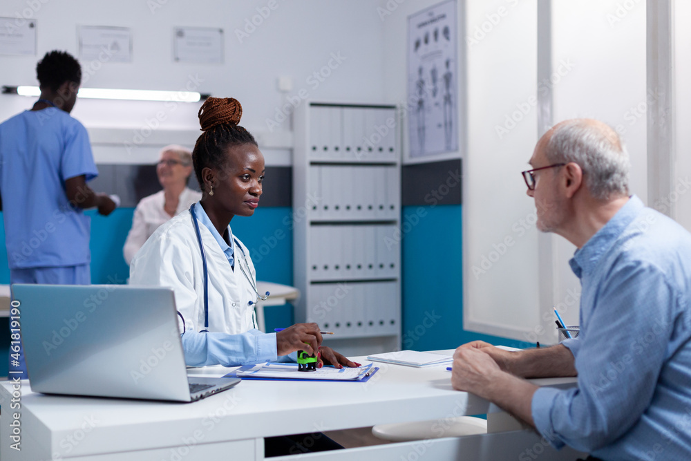 Woman of african ethnicity working as doctor discussing health checkup with elderly man at desk office. Black healthcare specialist doing professional consultation for old patient Woman of african ethnicity working as doctor discussing health checkup with elderly man at desk office. Black healthcare specialist doing professional consultation for old patient