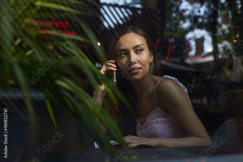 young asian pretty woman sitting at table of coffee shop, calling by smartphone. beautiful lady using cell phone shot through window glass outside of cafe. modern communication technology © Artem Zatsepilin