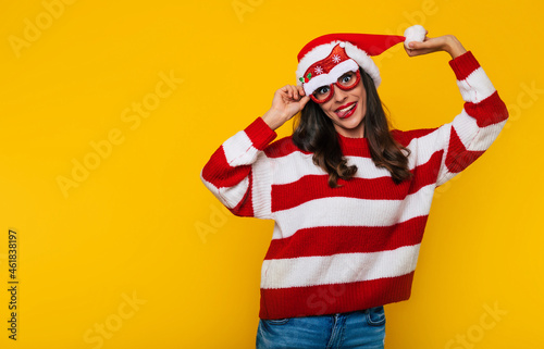Excited beautiful young woman in funny Christmas glasses and Santa hat is having fun while posing against a yellow wall