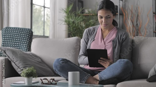 young arab woman sits on the sofa using tablet computer,mixed race brunette girl holding touchscreen device browsing internet onilne swipe screen indoors at home photo