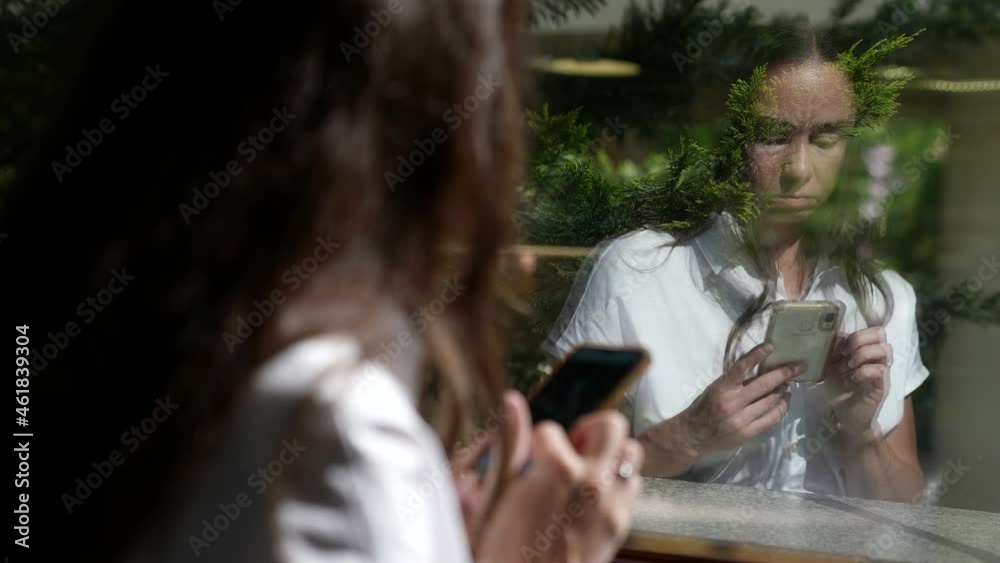a brunette with long hair, in a white dress and with a phone in hands is reflected in the window. over-the-shoulder view a brunette with long hair, in a white dress and with a phone in hands is reflected in the window. over-the-shoulder view