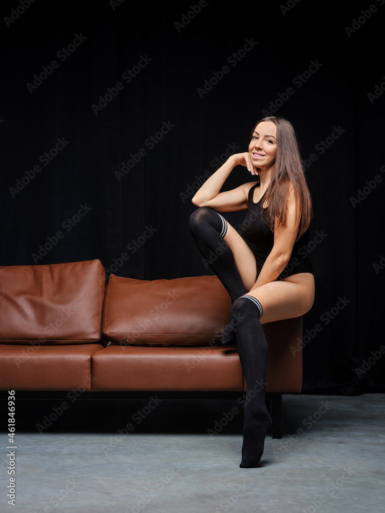 Middle age woman in bodysuit and overknee socks sits on armrest sofa