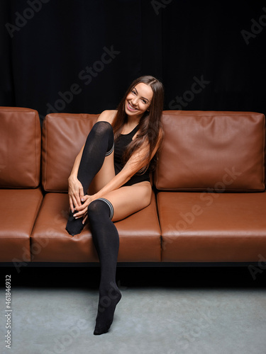 Middle age woman in bodysuit and overknee socks sits on leather sofa