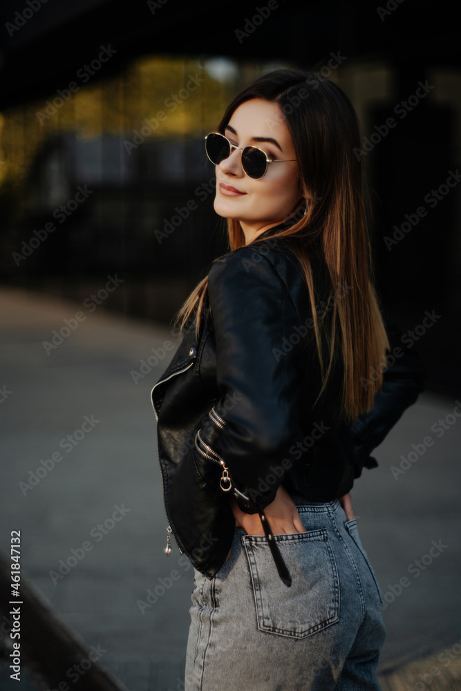 Fototapeta premium Young woman wearing black t-shirt, glasses and leather jacket posing against street