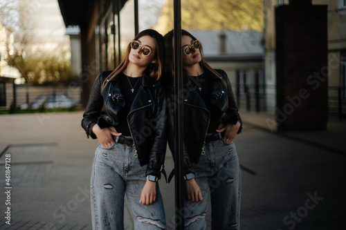 Fotografija Young woman wearing black t-shirt, glasses and leather jacket posing against str