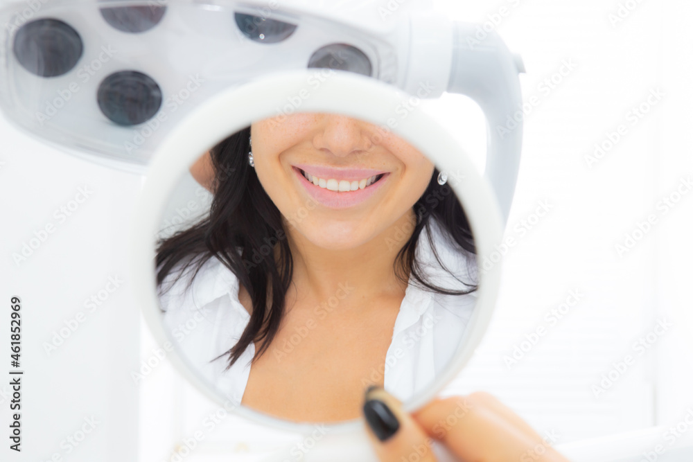 Beautiful woman looking in the mirror at the dentist office enjoying her beautiful smile and teeth after dental treatment at the dental clinic, selective focus Beautiful woman looking in the mirror at the dentist office enjoying her beautiful smile and teeth after dental treatment at the dental clinic, selective focus