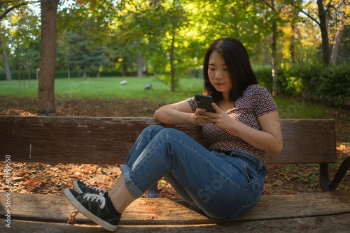 lifestyle portrait of young happy and beautiful Asian Chinese woman using internet app on mobile phone relaxed and cheerful at city park enjoying nature Autumn colors