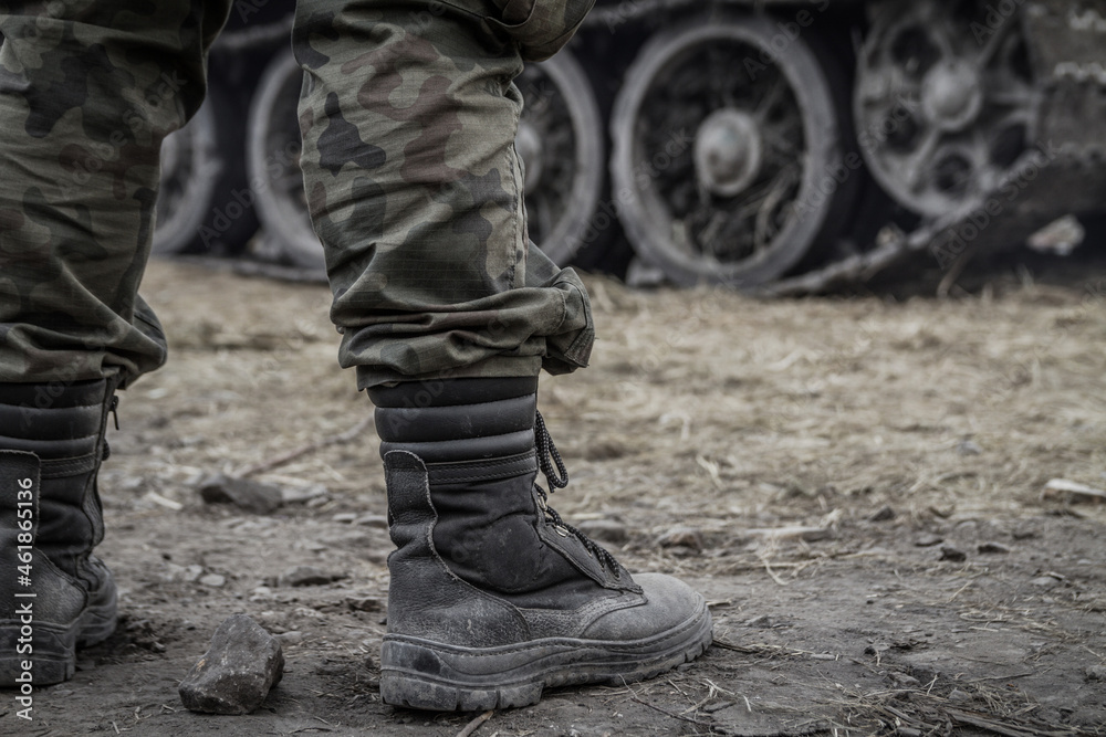 Close-up of soldier's legs on a battlefield, wearing military boots and woodland camo pants (camouflage trousers). With a tank tracked wheels in the background.