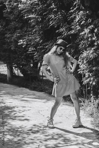 A girl in a summer dress and hat poses on the alley in the park while standing with her toes inward. Black and white photo