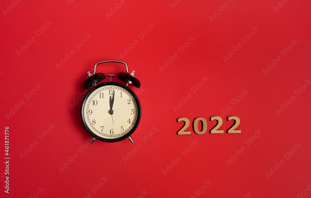 Top view of black alarm clock with midnight on the clock face and wooden numbers symbolizing the 2022 year. Christmas and New Year celebration concept over red background with copy space for ad Top view of black alarm clock with midnight on the clock face and wooden numbers symbolizing the 2022 year. Christmas and New Year celebration concept over red background with copy space for ad