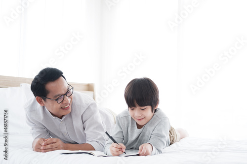 Smiling Asian father and son child is writing a book and doing homework together on bed in bedroom. Family and education on holiday concept.