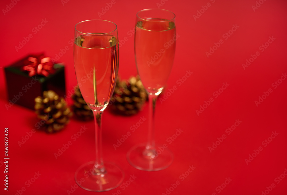 Two flutes of champagne or glasses with sparkling wine on the red background with pine cones ornate with golden paints and luxury Christmas gift in shiny green wrapping paper with glitter red bow Two flutes of champagne or glasses with sparkling wine on the red background with pine cones ornate with golden paints and luxury Christmas gift in shiny green wrapping paper with glitter red bow